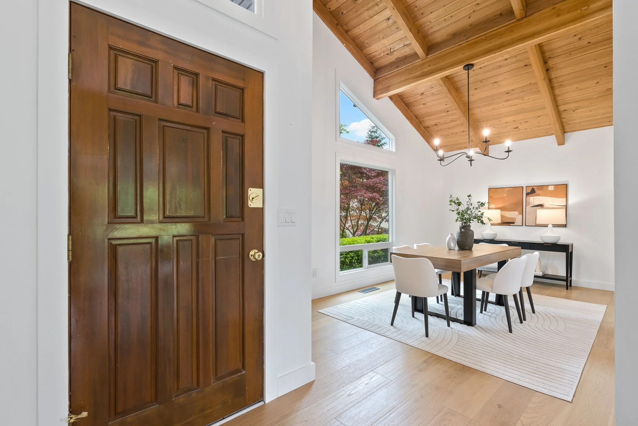 A dining room with a wooden table, six white chairs, a black sideboard, framed artwork, a chandelier, two large windows, and a wooden ceiling.
