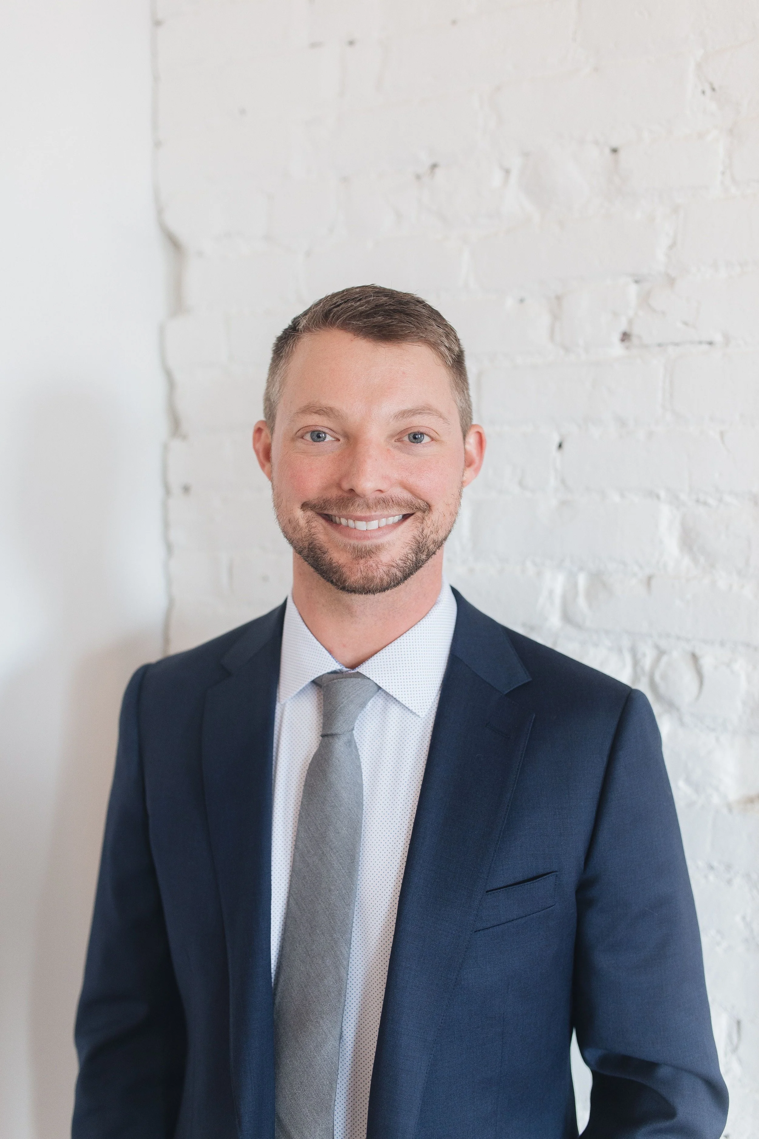 A man in a navy blue suit, white shirt, and gray tie, smiling and looking at the camera, standing against a white brick wall.