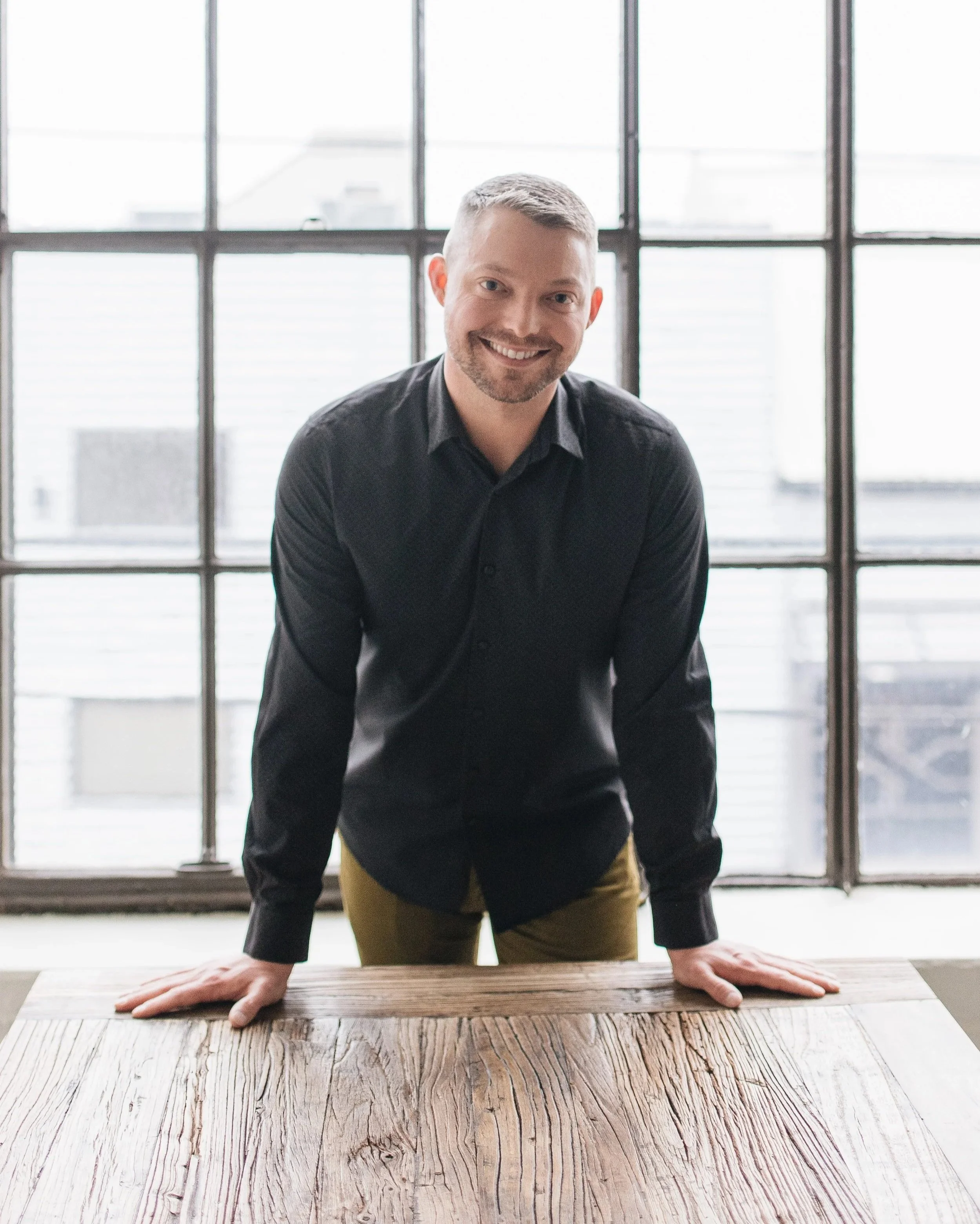 A man smiling and leaning forward with his hands on a wooden table in front of him, standing indoors near large windows with a cityscape view.