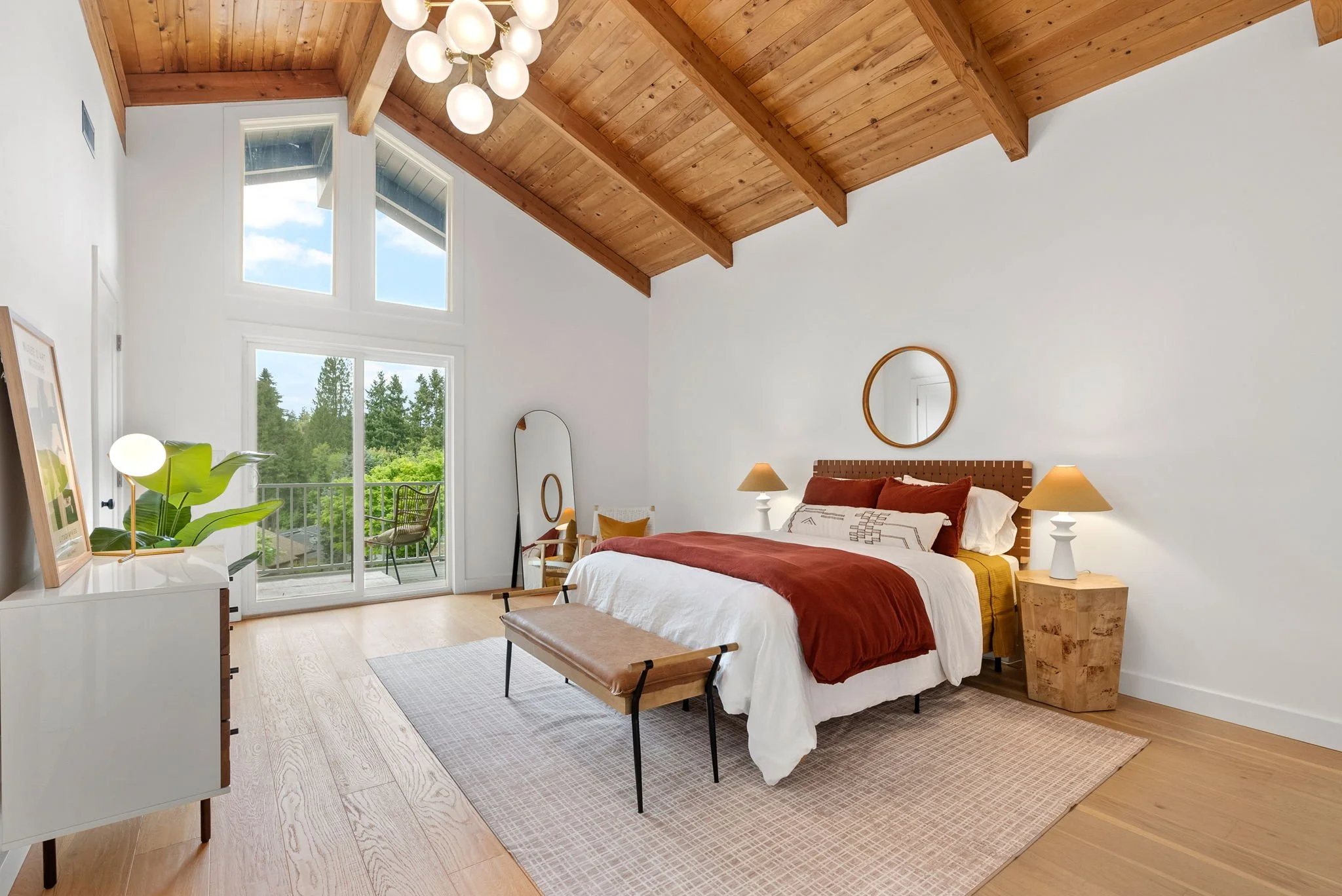 Bright bedroom with high vaulted wooden ceiling, large windows with view of trees, and a balcony. Bed with brown and white bedding, two nightstands with lamps, a bench at the foot of the bed, a mirror, and a dresser with a plant and artwork.