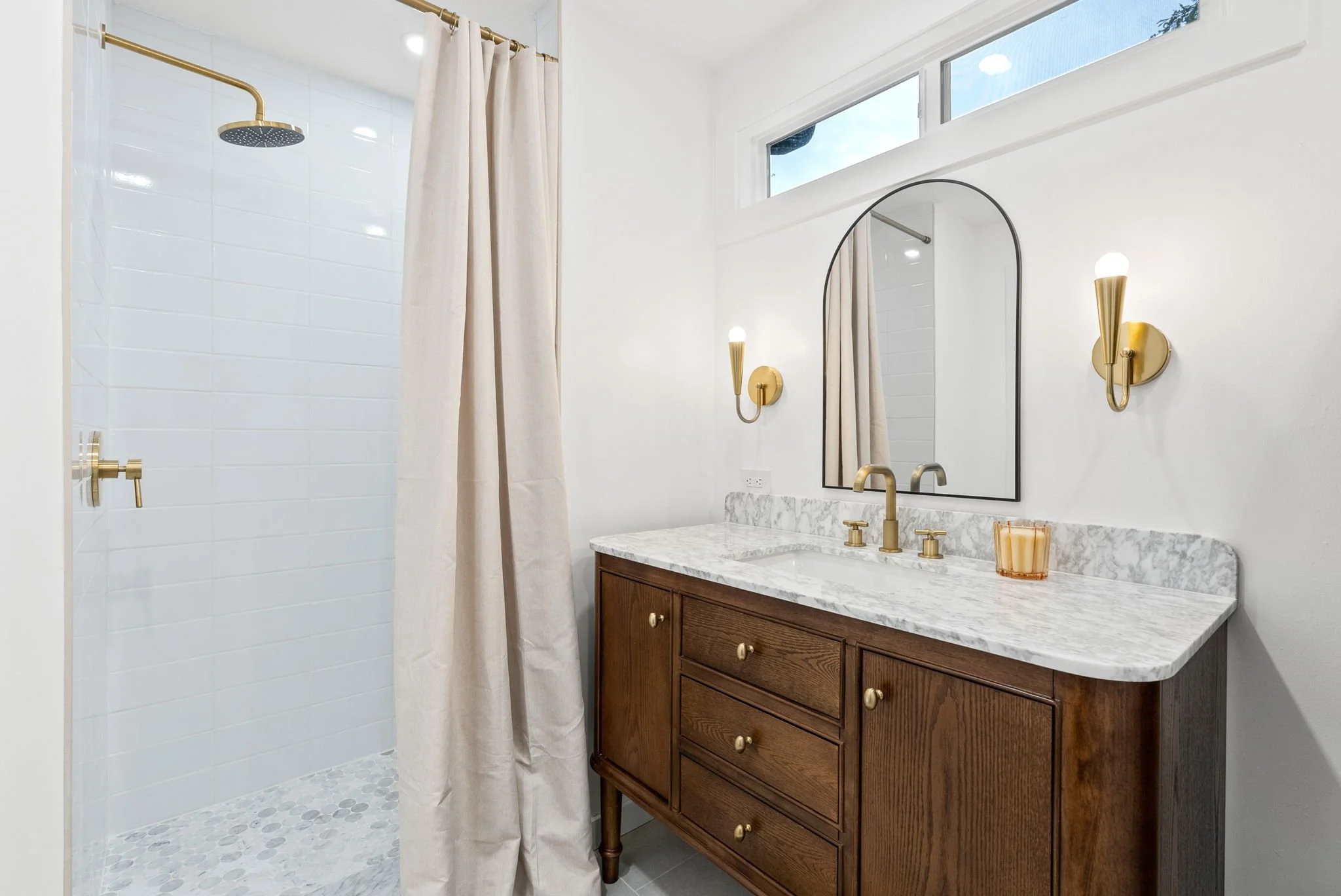 Bathroom with white walls, a marble countertop vanity with a wooden cabinet, a mirror, and gold fixtures. There is a shower with a shower curtain and an overhead showerhead.