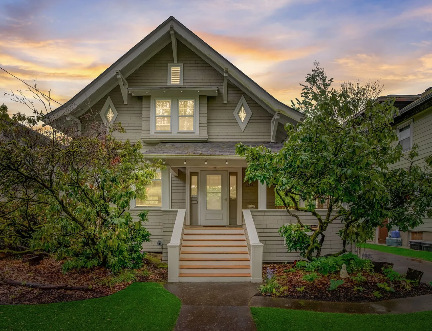 A two-story gray house with a front porch, steps leading up to the door, surrounded by green trees and a lush front lawn at sunset.