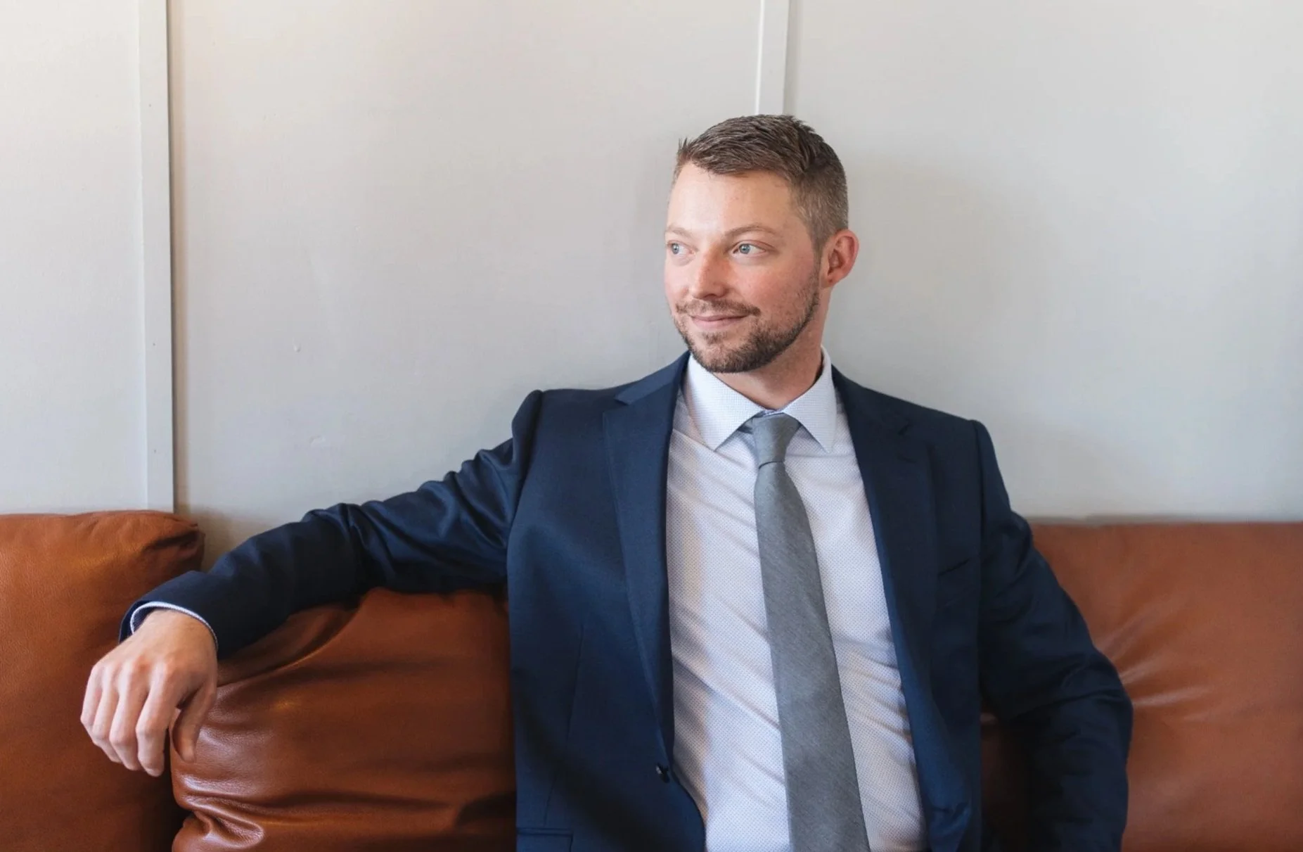 A man in a navy blue suit, white shirt, and gray tie sitting on a brown leather couch against a white wall, looking to his right with a slight smile.