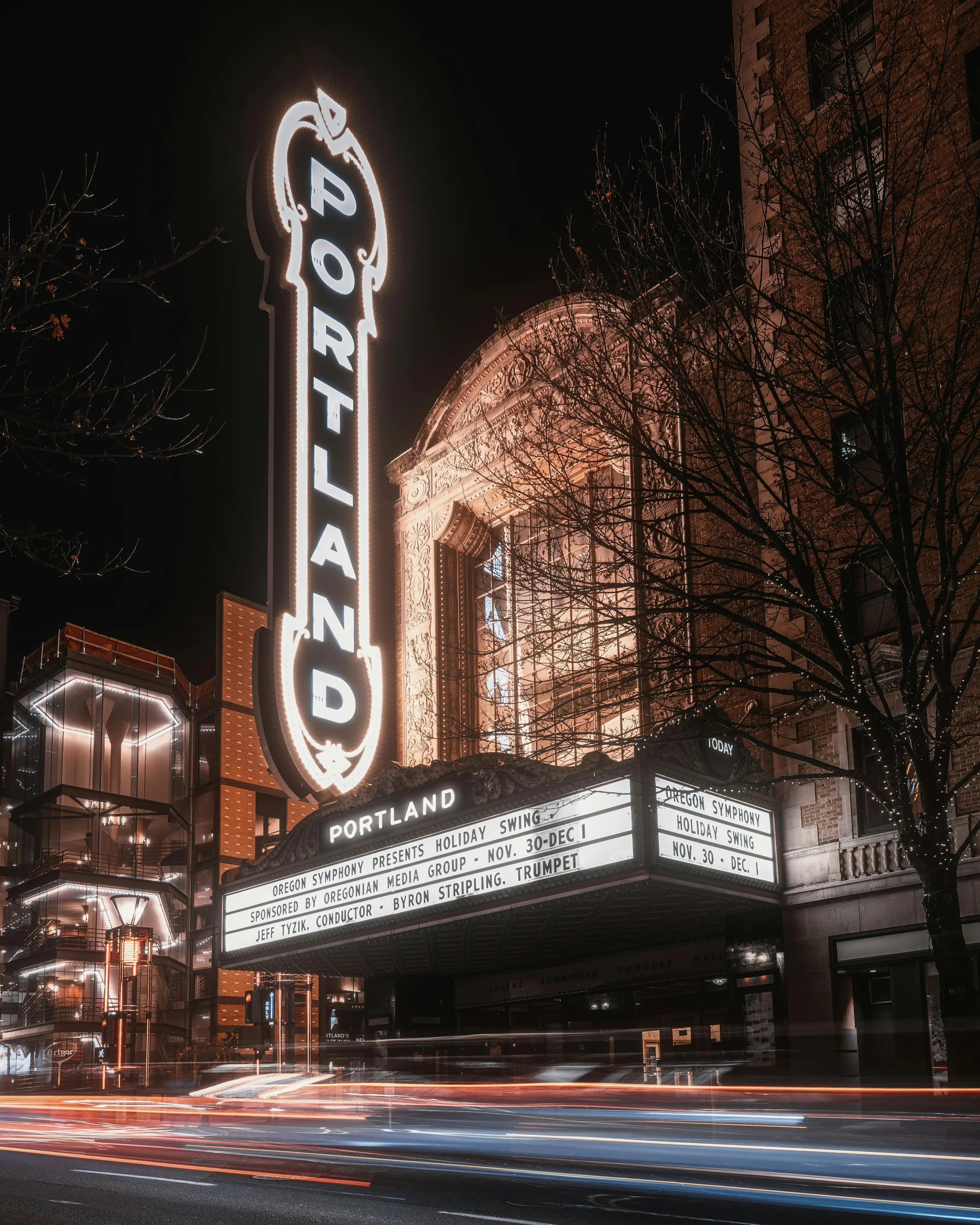 Night view of the Portland theater sign illuminated, with a marquee showing a holiday swing concert by the Oregon Symphony and other details. The theater's historic facade is adorned with detailed architectural design, and a tree with string lights is visible in front.