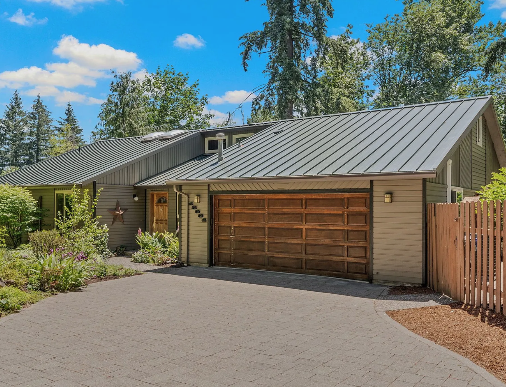 Front view of a house with a metal roof, a wooden garage door, and a side entrance door, surrounded by landscaping and trees under a bright blue sky.