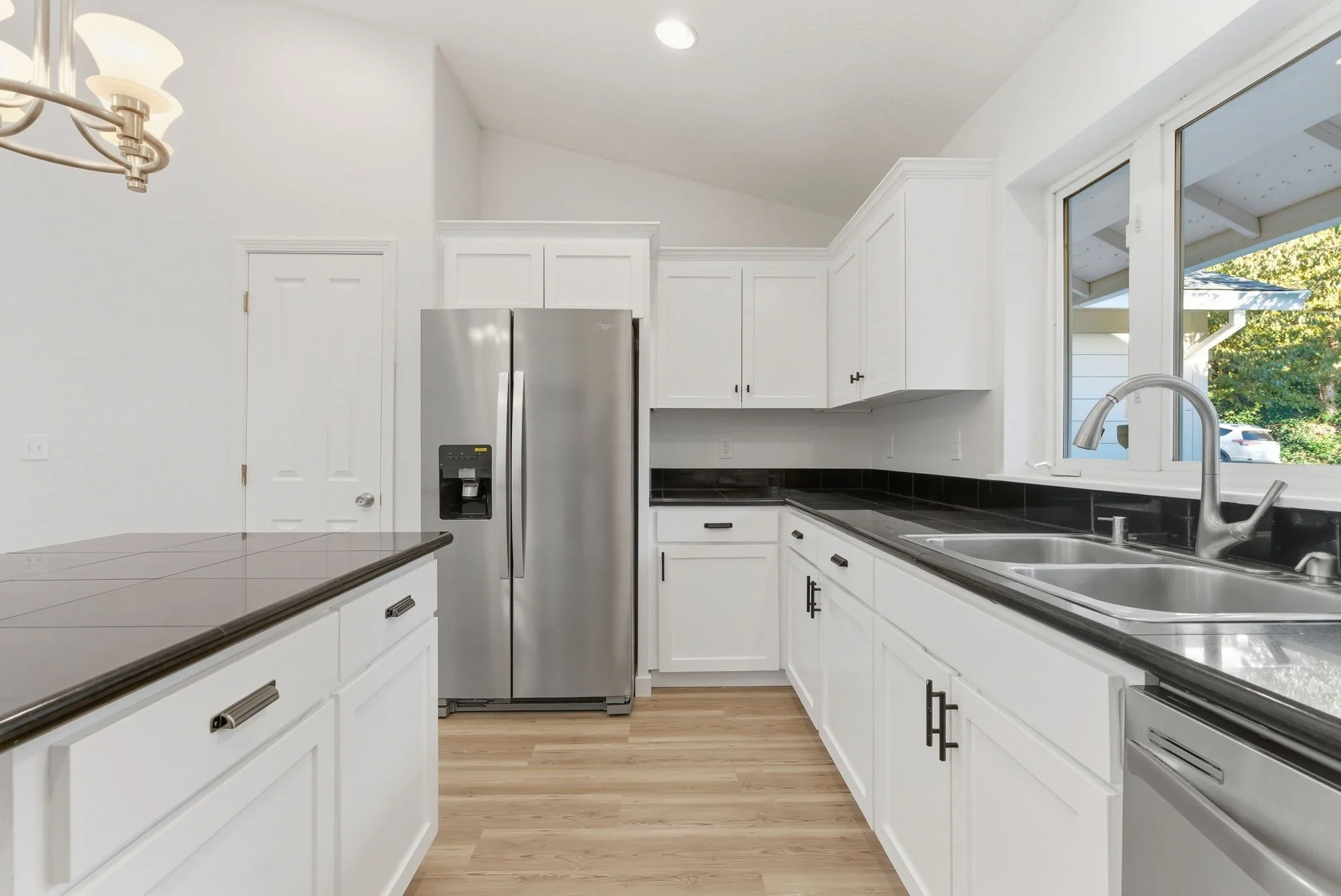 Kitchen with white cabinets, stainless steel refrigerator, black countertop, double kitchen sink, and window overlooking outdoor greenery.