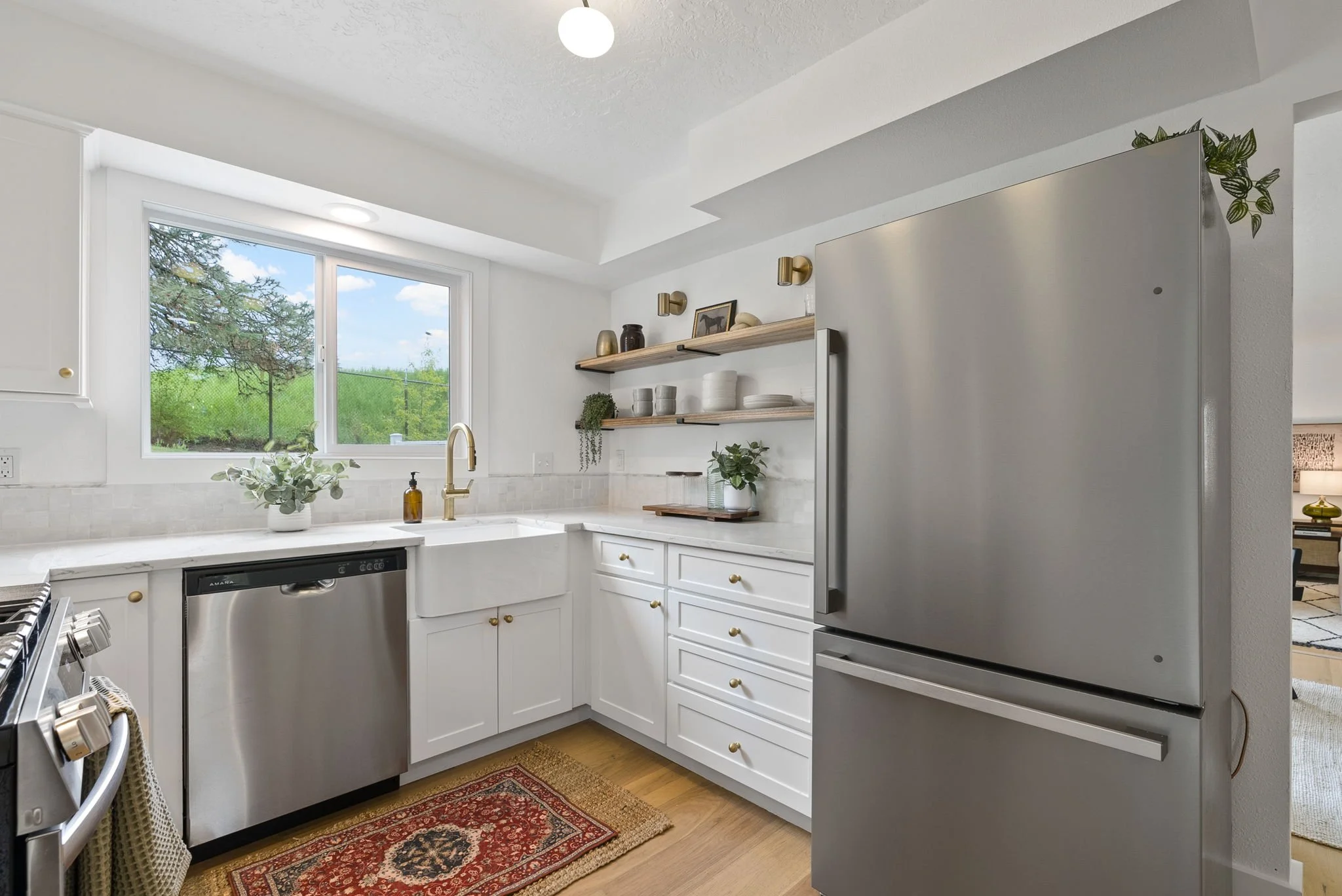 Modern white kitchen with stainless steel appliances, a window with a view of trees, open wooden shelves with dishes and decor, a green plant, and a small decorative rug.