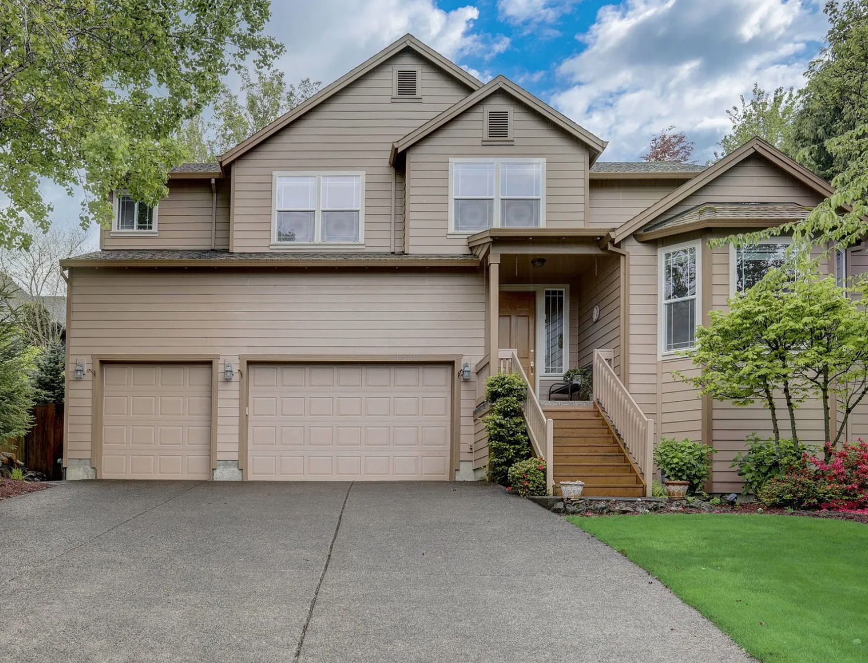 A two-story beige house with a double garage, front porch, wooden stairs, and landscaped yard with green grass, small trees, and bushes under a partly cloudy sky.
