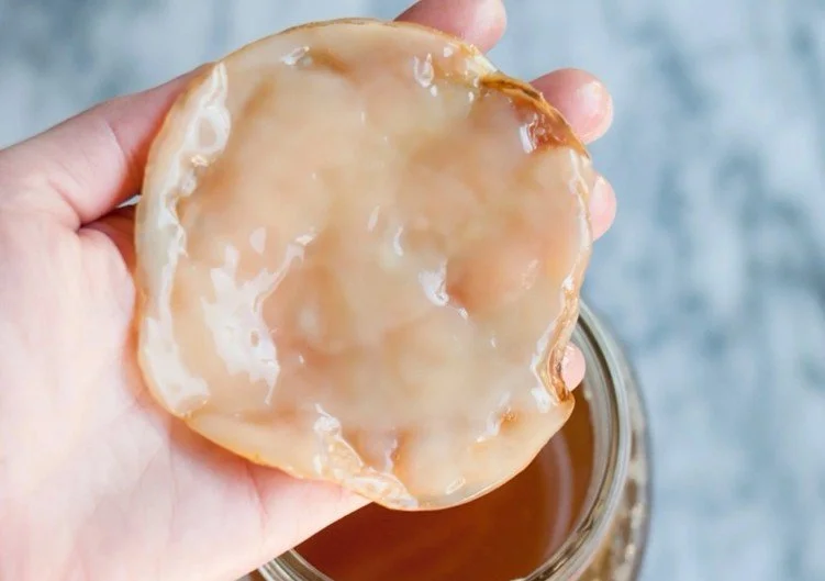 A hand holding a glazed donut over a jar of caramel sauce.