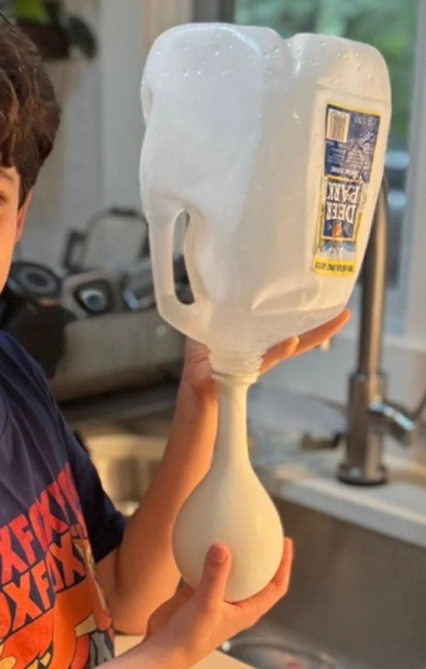 A person holds a plastic container filled with a frothy, beverage-like substance, attached to a pitcher with a narrow neck, in a kitchen setting.