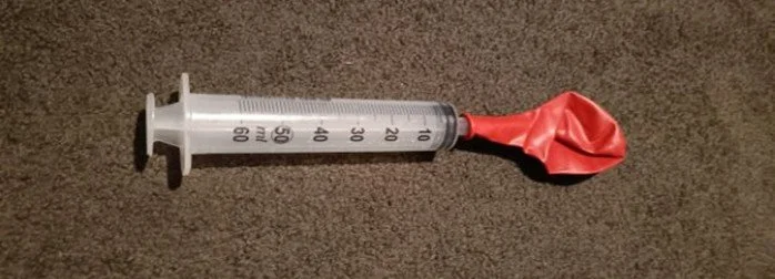 A syringe with a deflated red balloon attached to the needle end, lying on a brown surface.