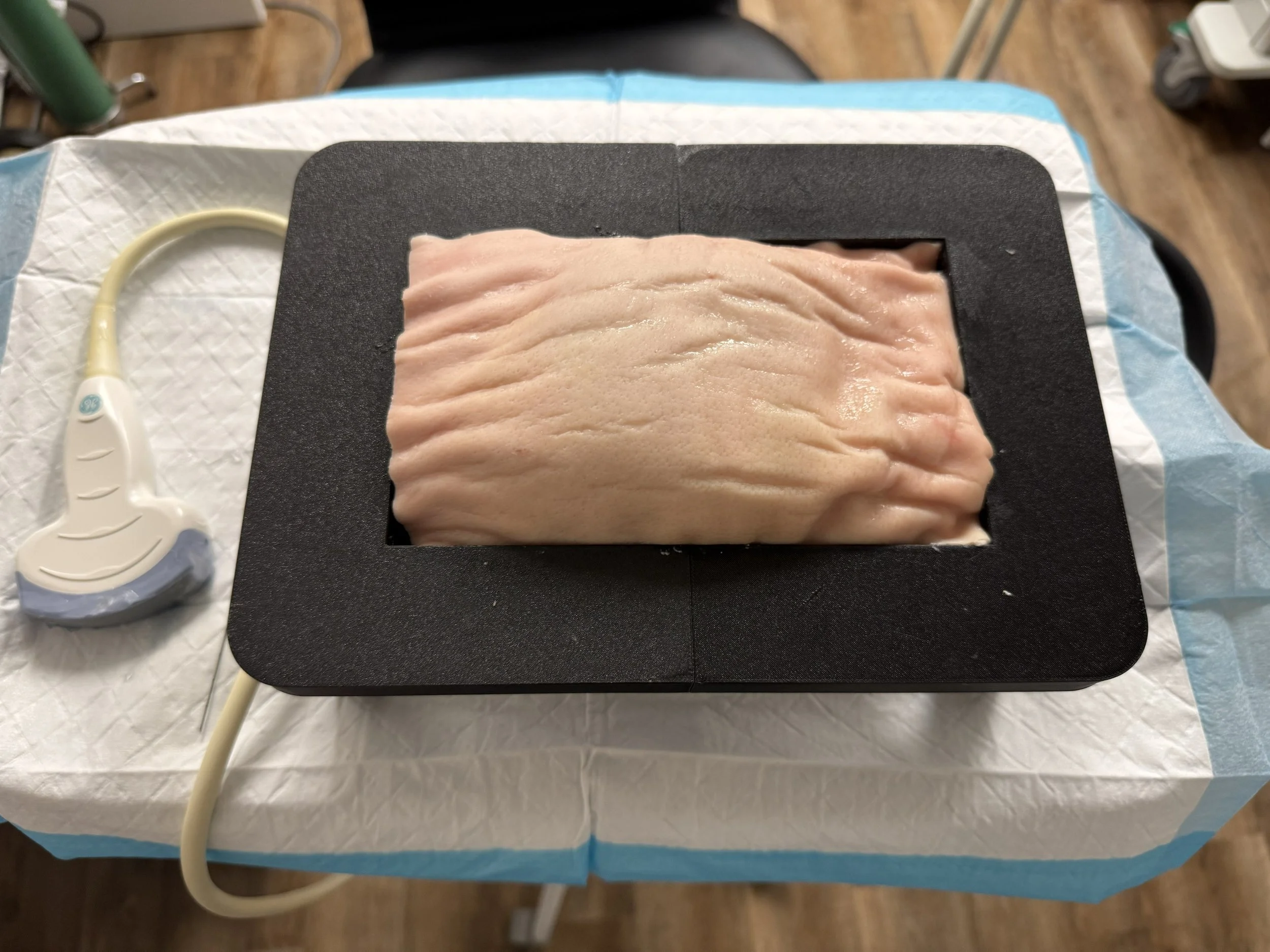 Medical ultrasound probe connected to an ultrasound device, placed on a draped surface beside a black rectangular X-ray or imaging platform with a large, folded medical specimen or tissue sample on top.