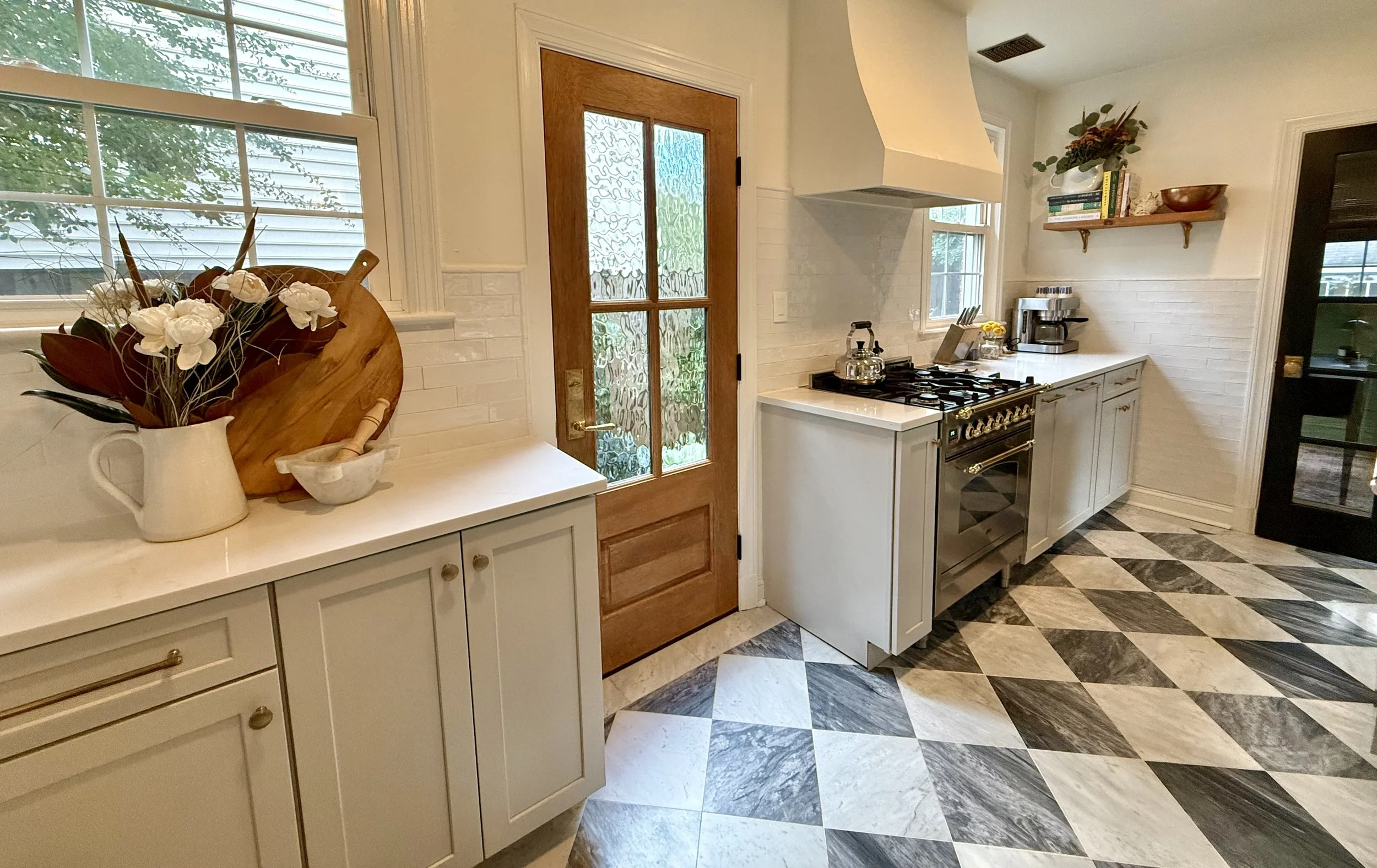 A bright North Virginia kitchen with neutral cabinetry, a gas stove, a shelf with books and decorative items, a door with glass panels, and a window, featuring marble checkered flooring.
