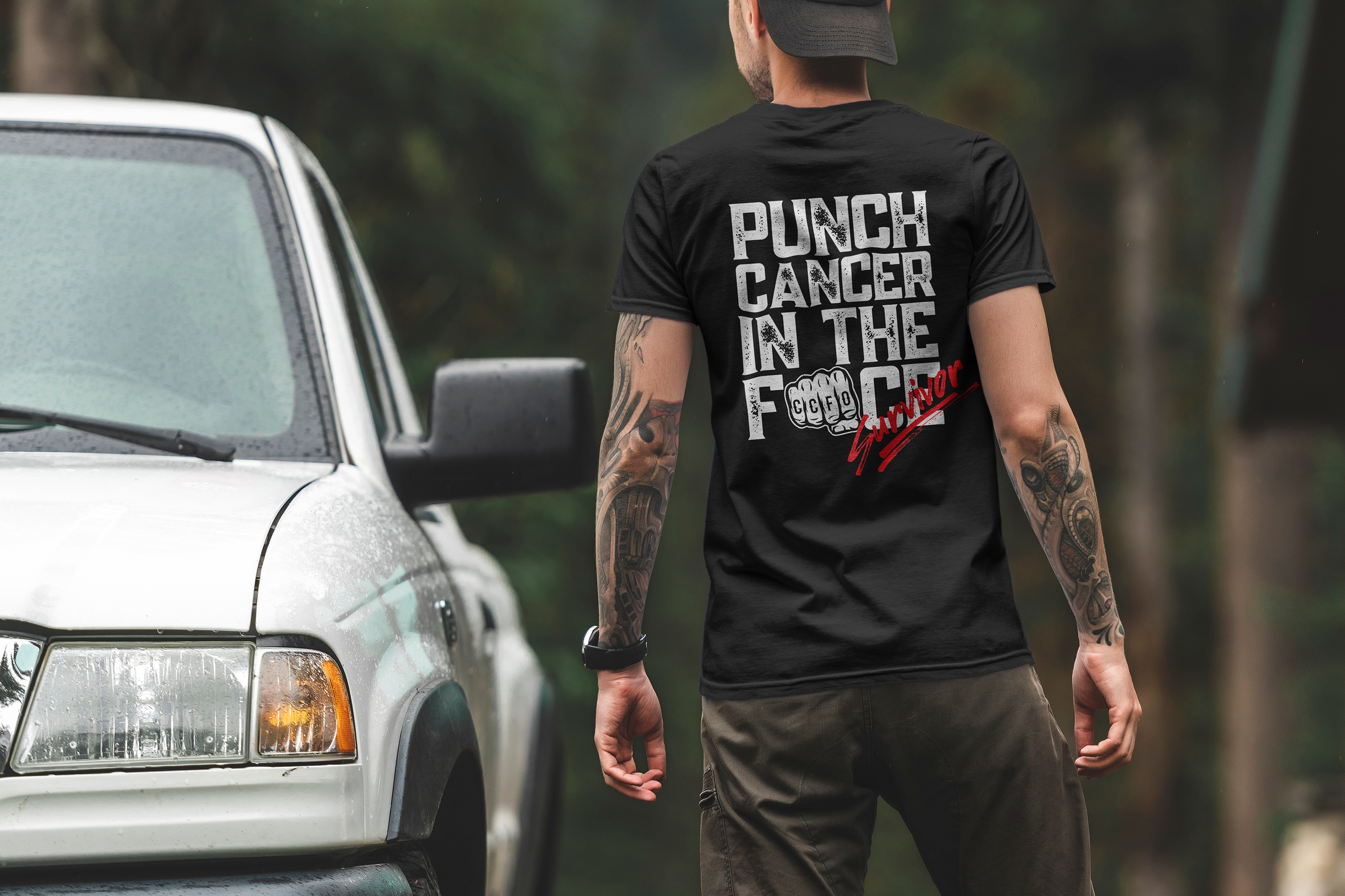 A man with tattoos on his arms is standing next to a white vehicle with foggy windows. He is wearing a black t-shirt with the text 'PUNCH CANCER IN THE FACE' and a red 'Survivor' script on it, and a black cap.