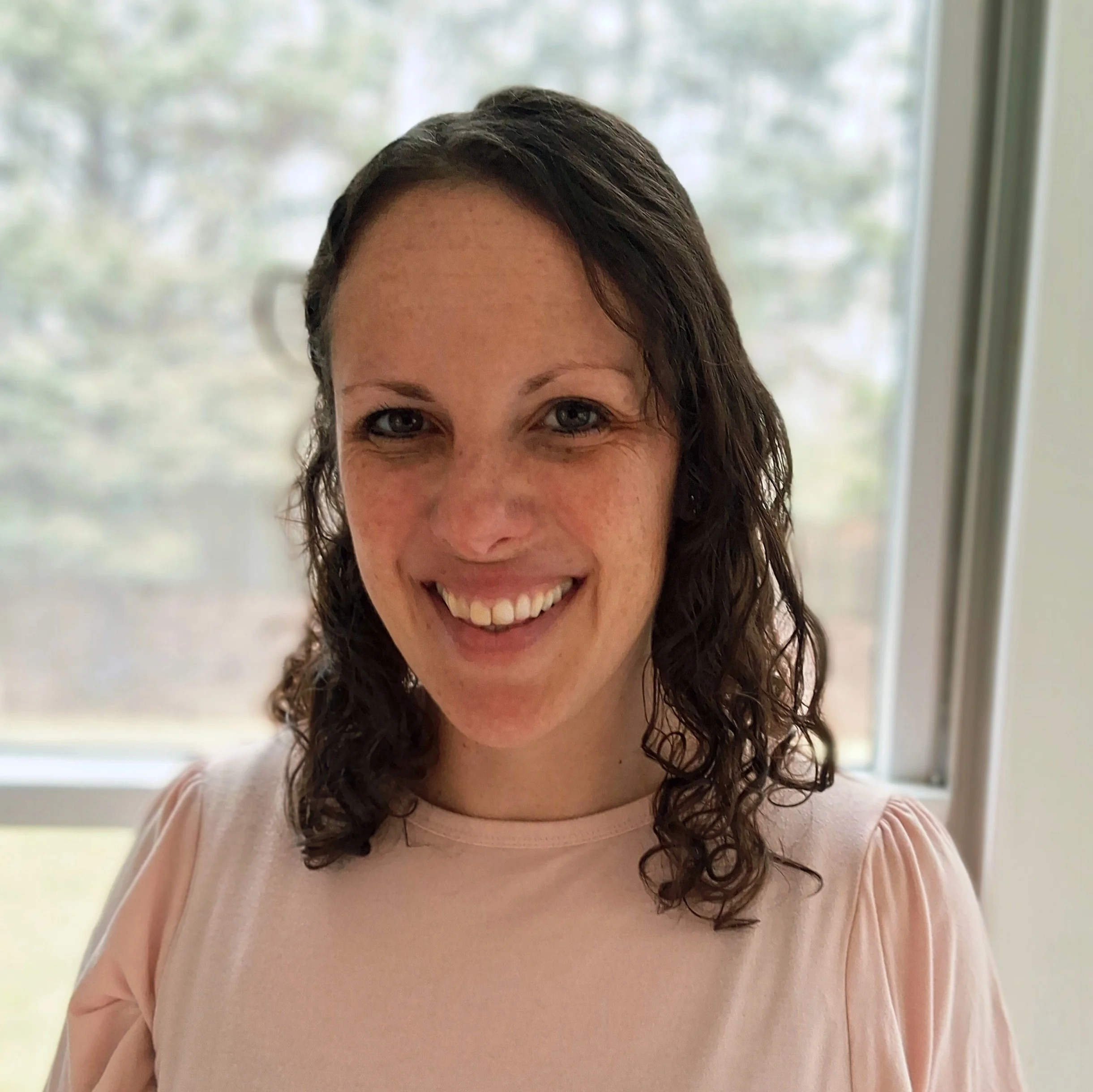 A woman with curly brown hair smiling, wearing a light pink top, standing near a window with a blurred outdoor background.