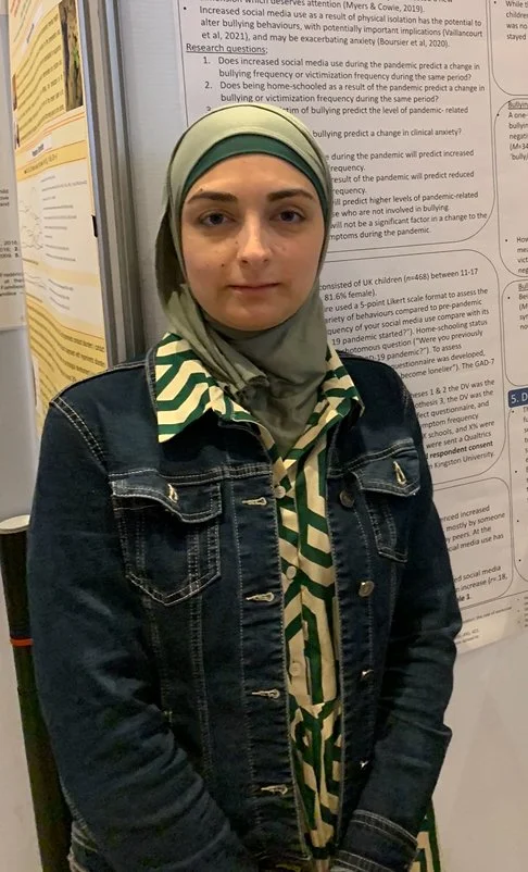 A young woman wearing a light green hijab with gray, green, and white patterns, and a denim jacket standing in front of a scientific research poster