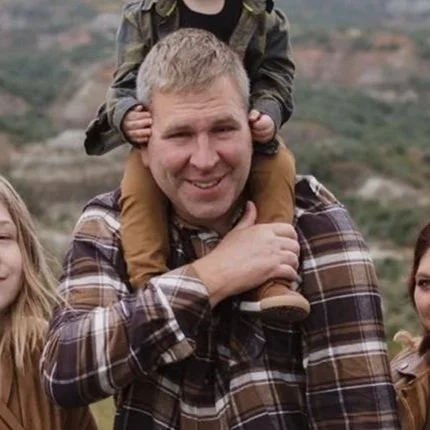 A man smiling with a young child sitting on his shoulders during an outdoor outing