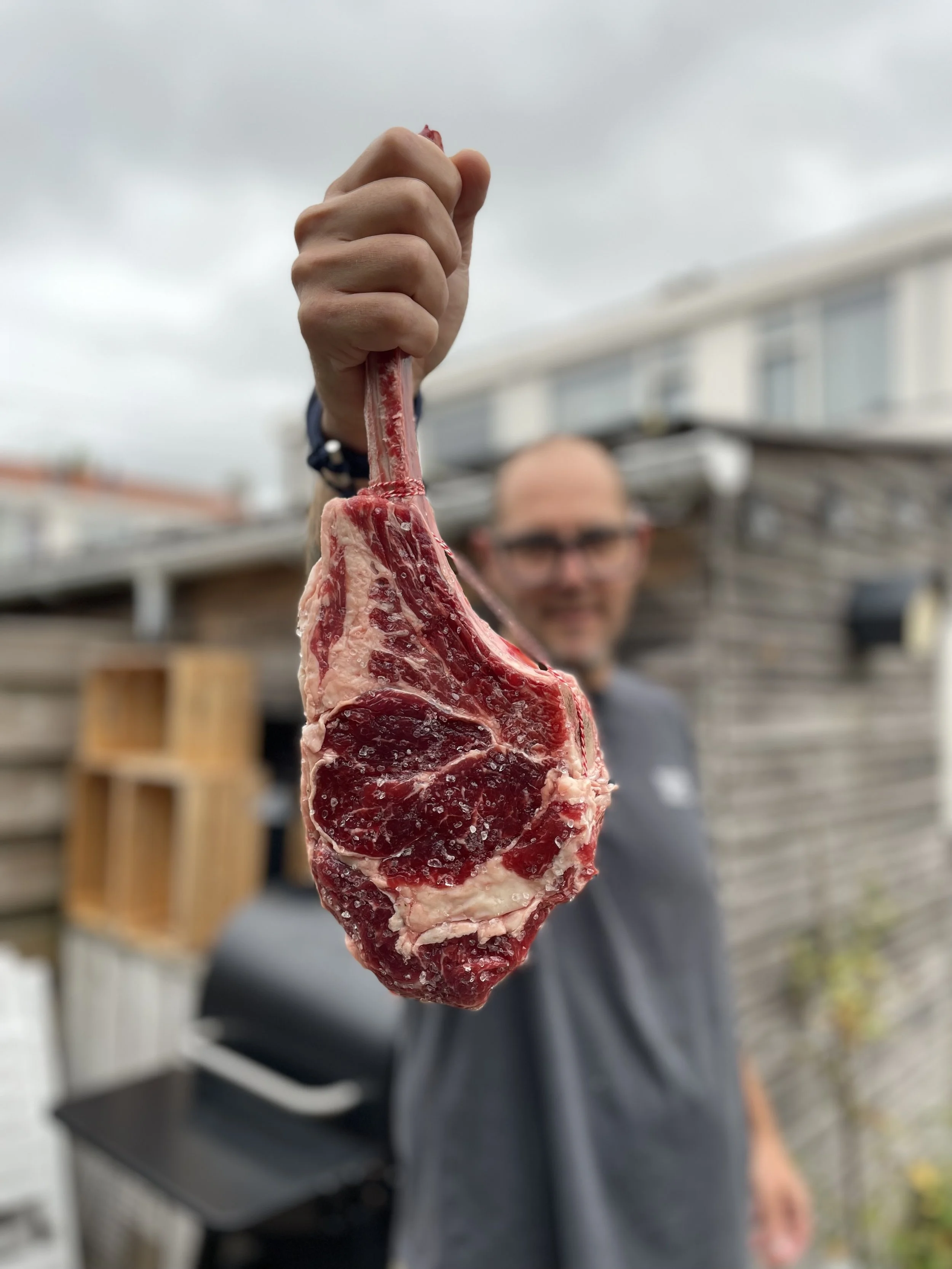 Person holding a large raw bone-in steak towards the camera outdoors, with a blurred background of a person, a wooden fence, and a cloudy sky.