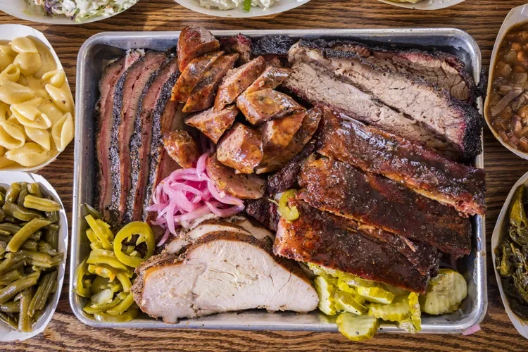 Een grote metalbak met verschillende soorten BBQ vlees zoals brisket, pulled pork, worsten, en geroosterd vlees, omringd door bijgerechten zoals groene bonen, pasta, en zuurkool.