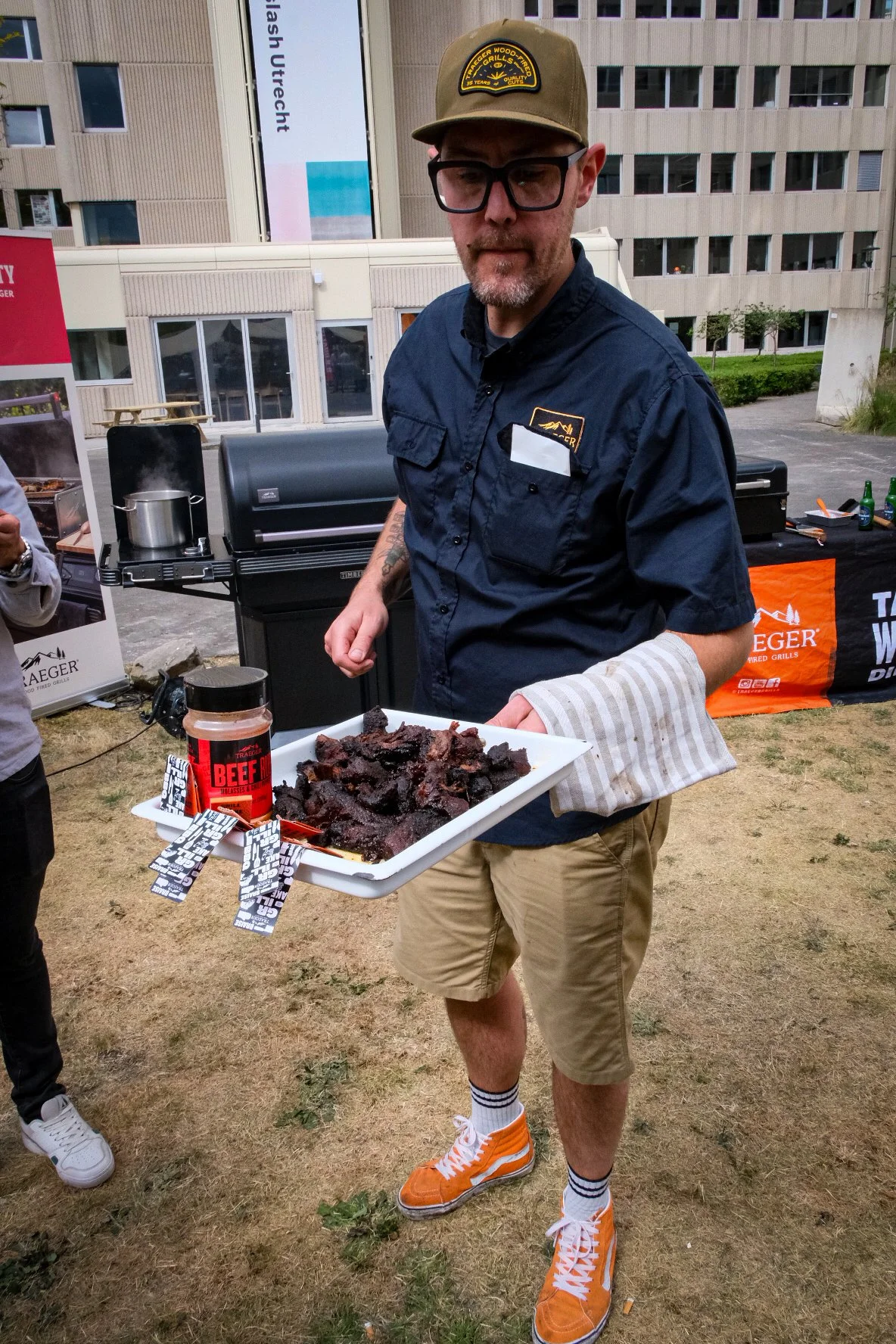 A man holding a tray with cooked barbecue meat, standing outdoors near a barbecue grill, wearing glasses, a brown hat, a navy blue shirt, beige shorts, and orange sneakers.