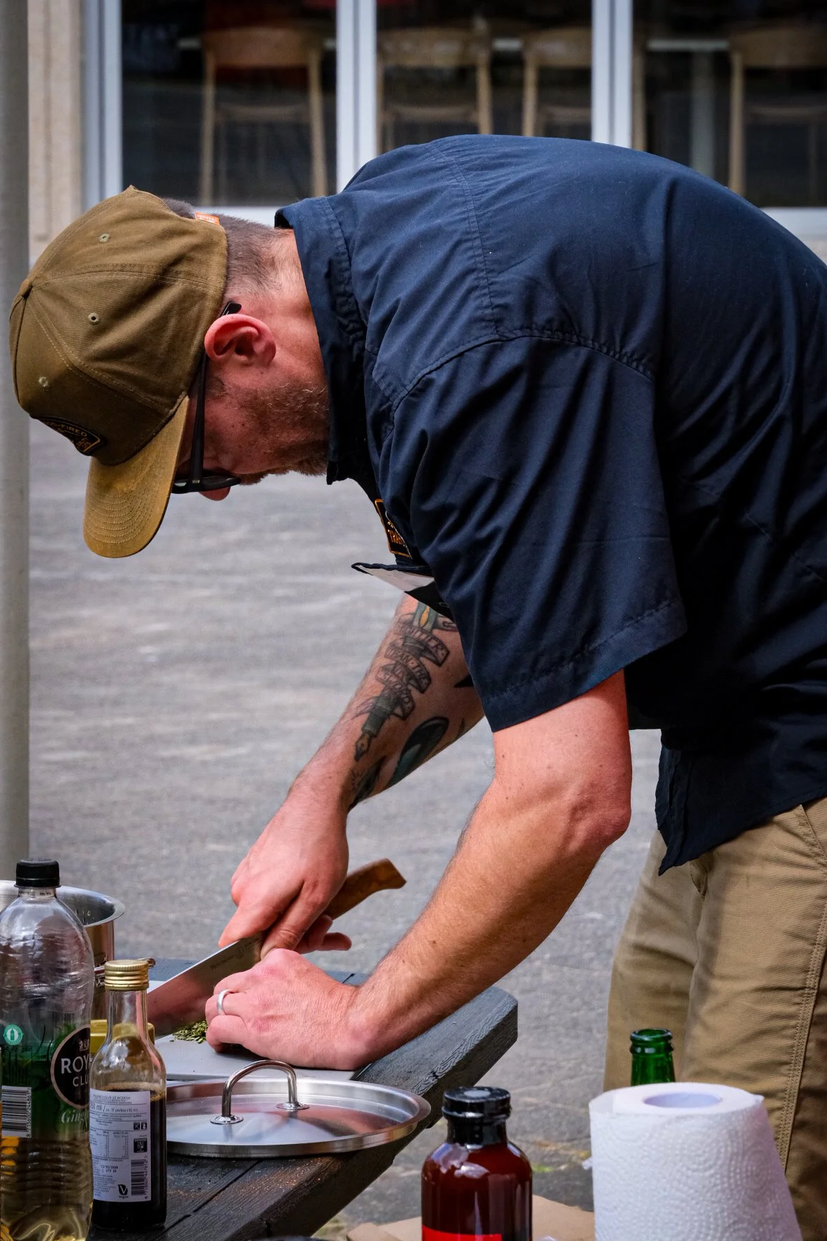 Man in een blauwe uniform en een groene pet snijdt iets met een grote keukenmes op een tafel, buiten.