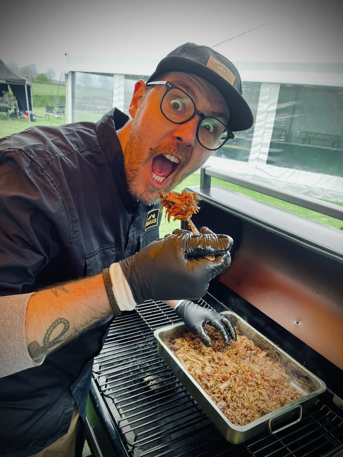 Man met zwarte bril en hoed grilt en eet gekruid vlees uit een braadblik onder een tent, met een groene buitenomgeving op de achtergrond.