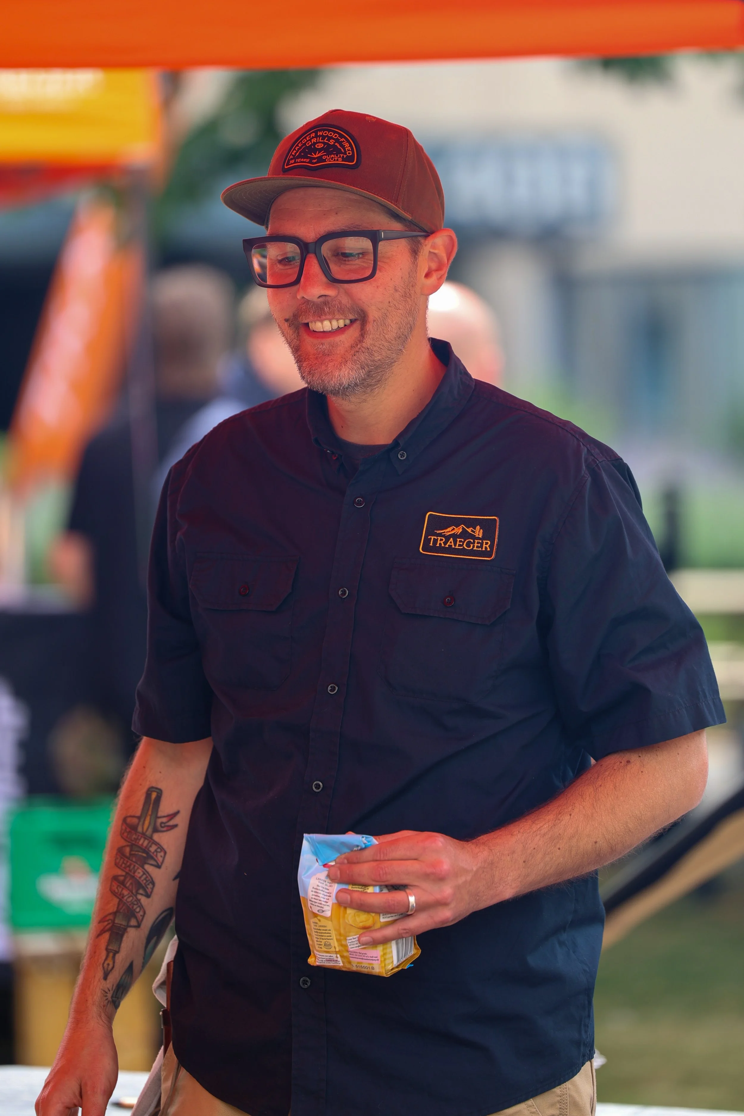 Man met een grijze baard en bril, lachend, draagt een zwart overhemd met een 'Traeger'-logo en een rood petje, houdt een zakje chips, tatoeage op zijn linker onderarm.