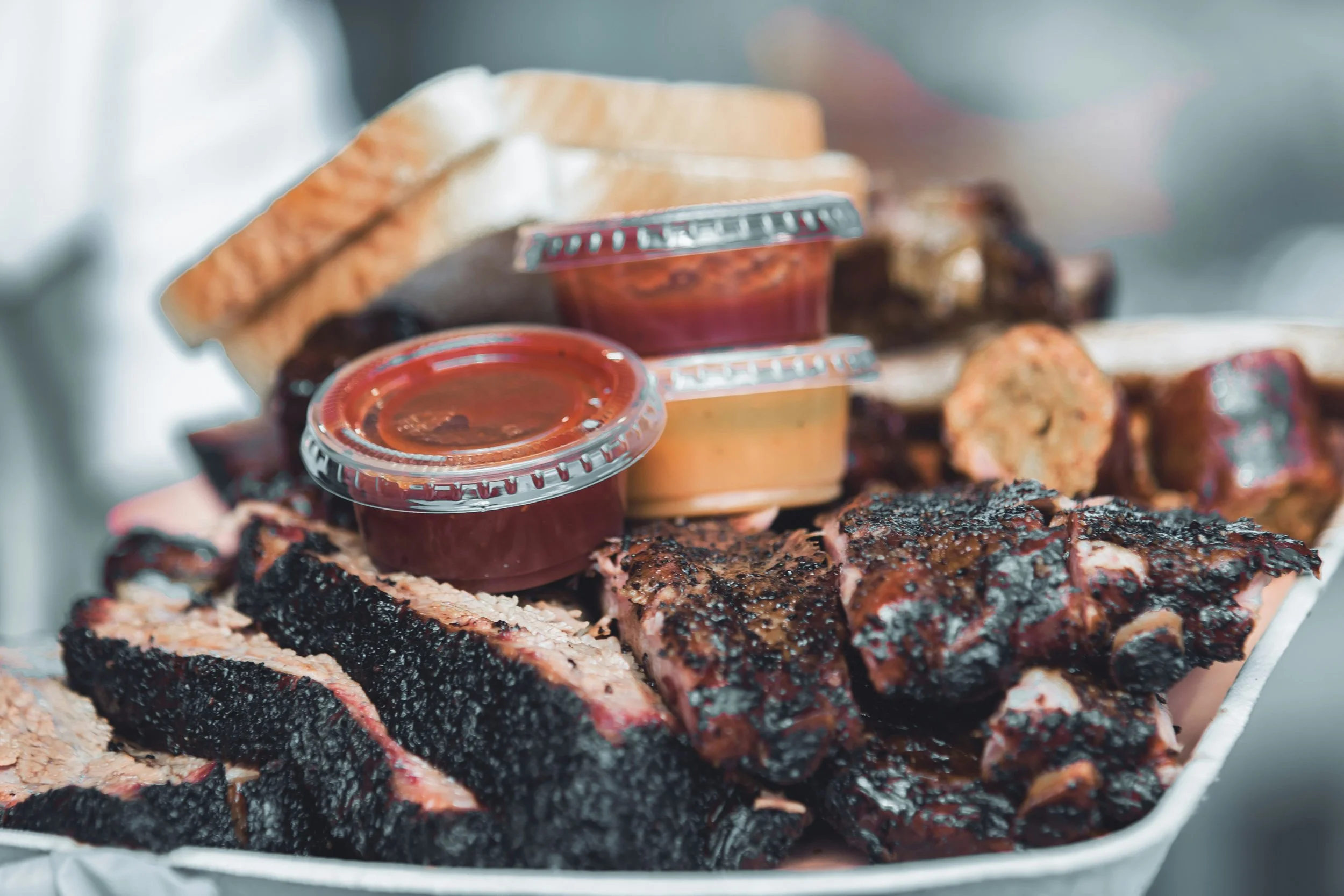 Tray of barbecue food with ribs, sausage, bread, and sauces.