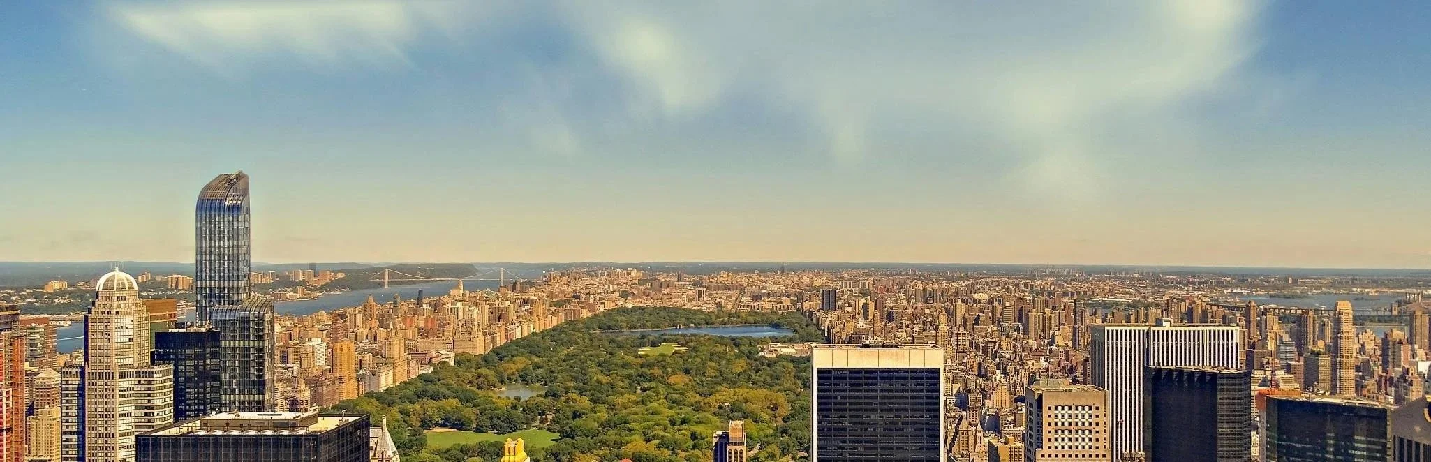 A panoramic view of New York City with Central Park in the foreground, tall skyscrapers, and bridges over rivers under a partly cloudy sky during daytime.