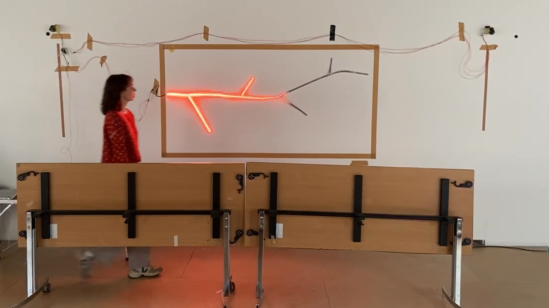 A girl stands in front of a framed art installation with a branching light pattern resembling a tree in a gallery or classroom setting.