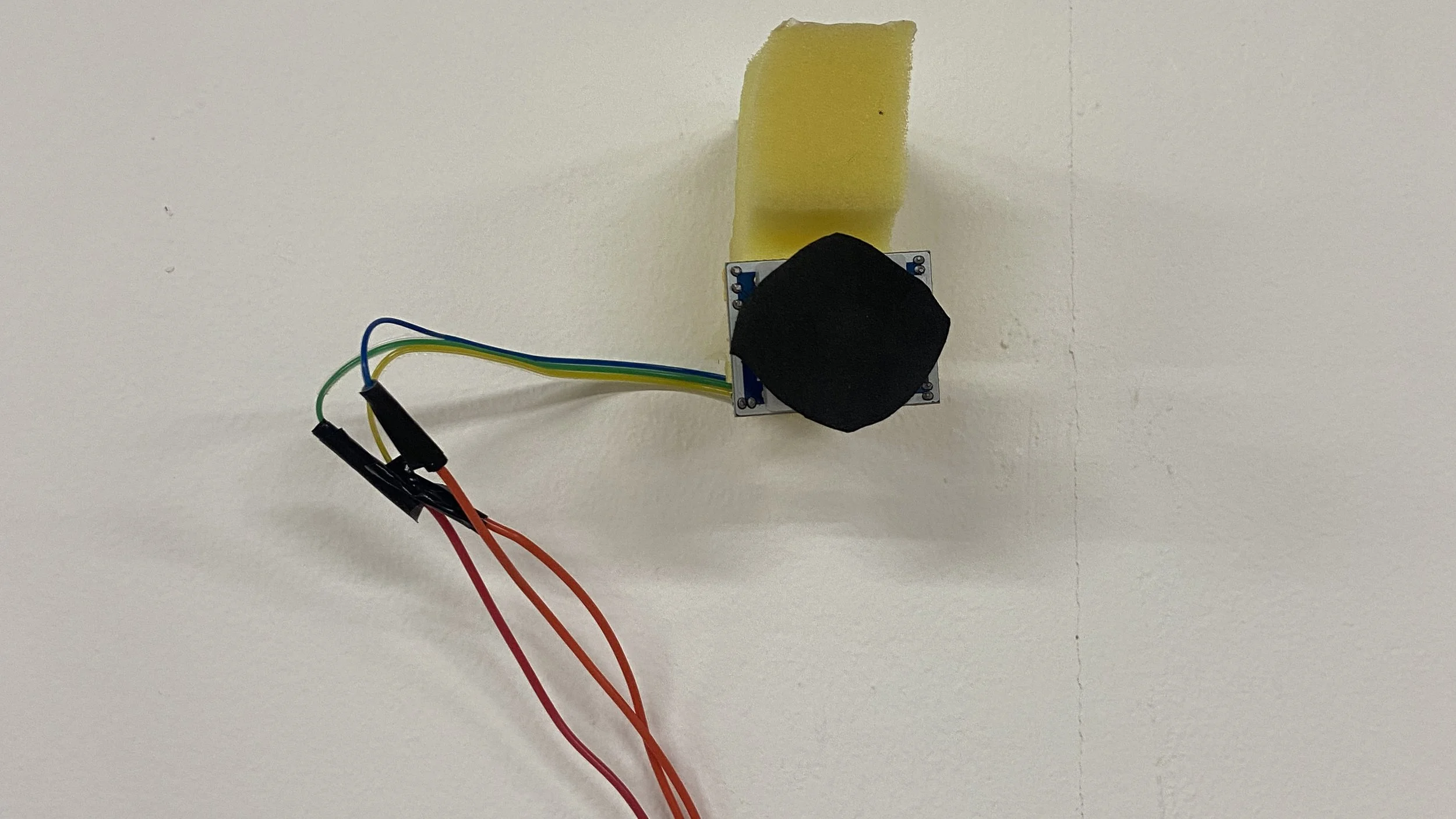 Electronic PiR sensor mounted on a wall with wires connected, including a yellow foam piece and a black square component attached to a small circuit board.