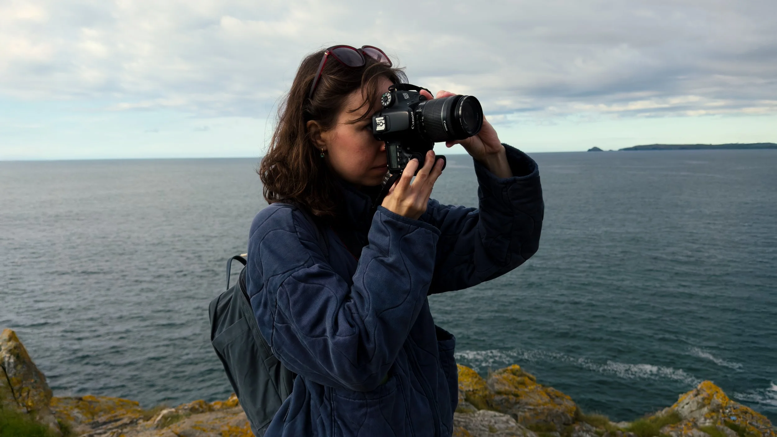 A woman with brown hair, sunglasses on her head, dressed in a blue jacket, taking a photograph with a camera near a body of water with rocky terrain and distant islands under cloudy skies.