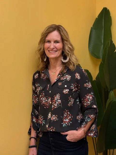 A smiling woman with curly blond hair, wearing a black floral blouse, jean pants, large earrings, and a necklace, standing with her hand in her pocket against a yellow wall with large green leaves.
