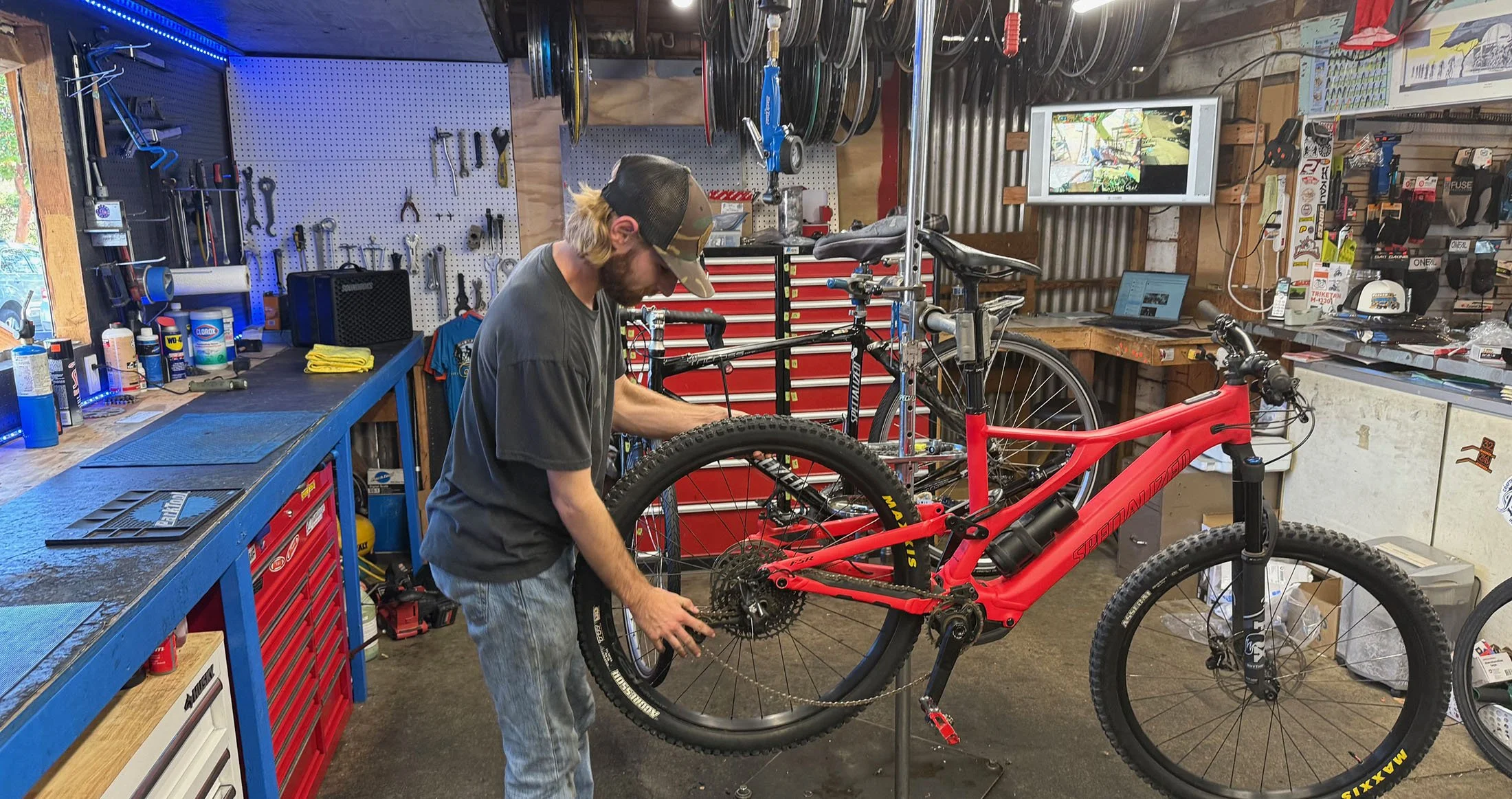 A man working on repairing a red mountain bike in a well-organized bike repair shop. The shop has tools hanging on a pegboard, a workbench with various supplies, and a TV screen on the wall displaying a bike trail.