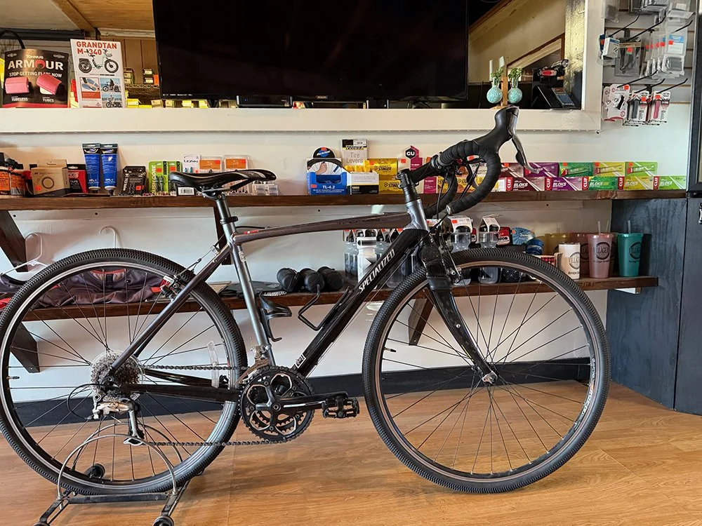 A black and silver Specialized road bike with drop handlebars, positioned on a bike stand in a store with shelves of bike accessories and supplies behind it.