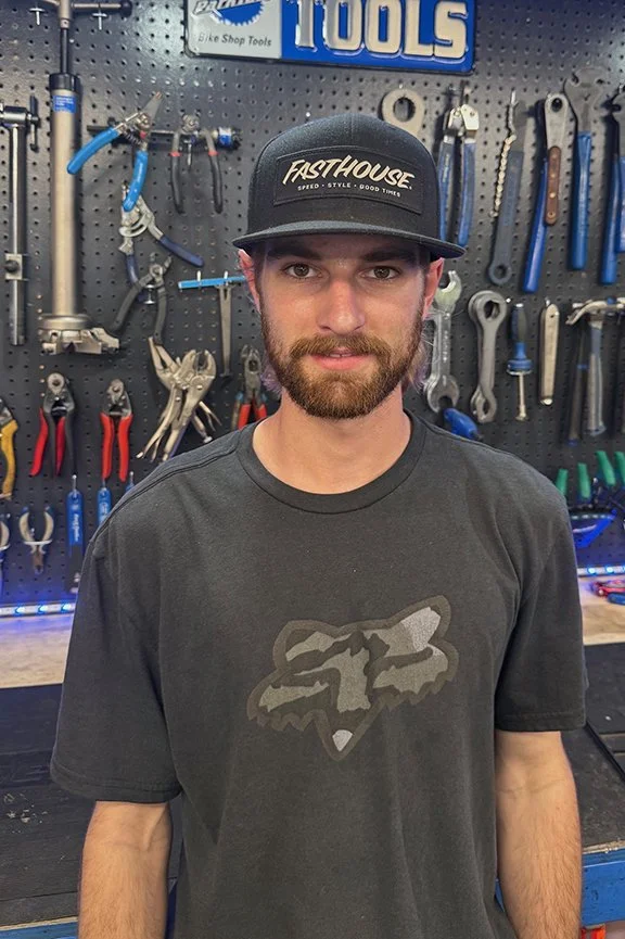 A young man with a beard and mustache wearing a black cap with 'Fasthouse' logo and a dark gray T-shirt with a fox head bold design, standing in a workshop with various tools hanging on a black pegboard wall behind him.