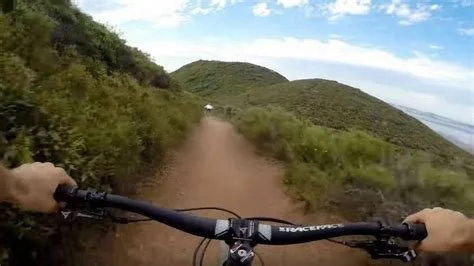 First-person view of a mountain biker riding on a dirt trail through green hills under a partly cloudy sky.