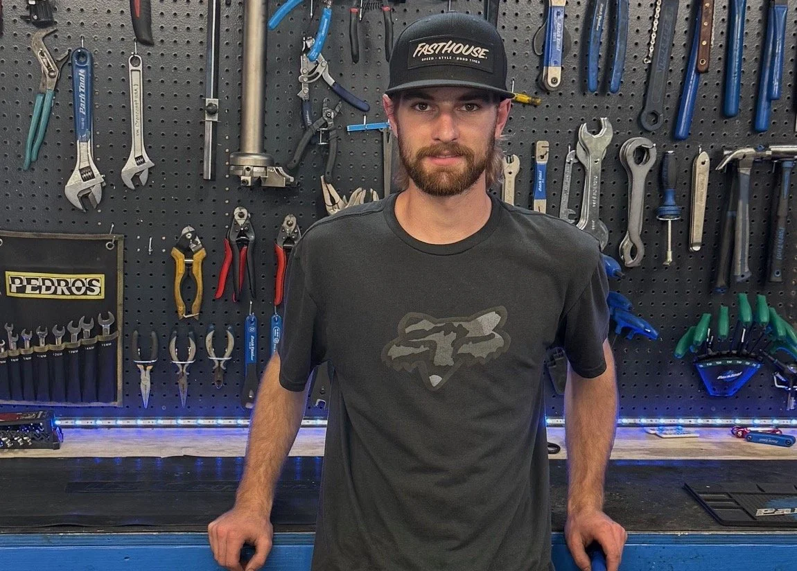 A man with a beard and mustache wearing a black baseball cap and gray Fox Racing T-shirt standing in front of a tool wall with various hand tools hanging on a black pegboard.