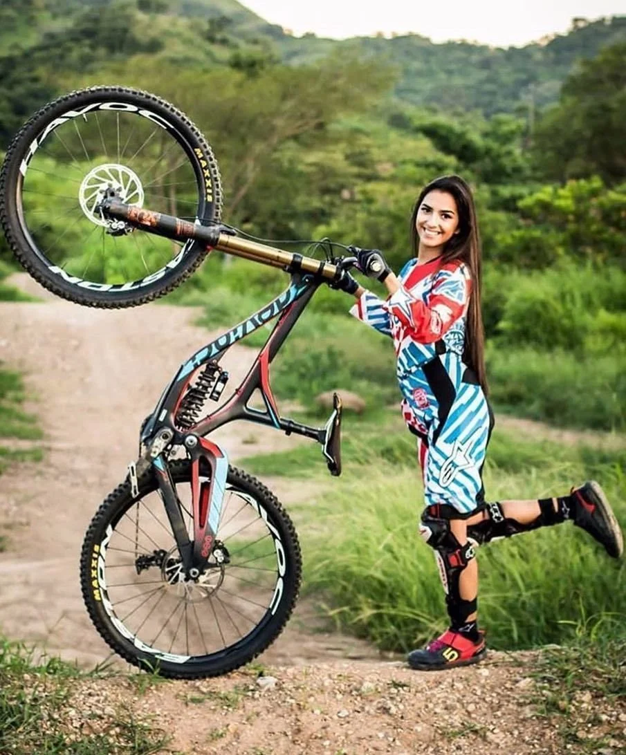 A woman with long brown hair, wearing a blue, red, and white motocross outfit, is holding an orange and black mountain bike with one wheel resting on the ground and the other wheel lifted in the air. She is standing on a dirt trail in a green, hilly outdoor area with trees and grass, smiling at the camera.