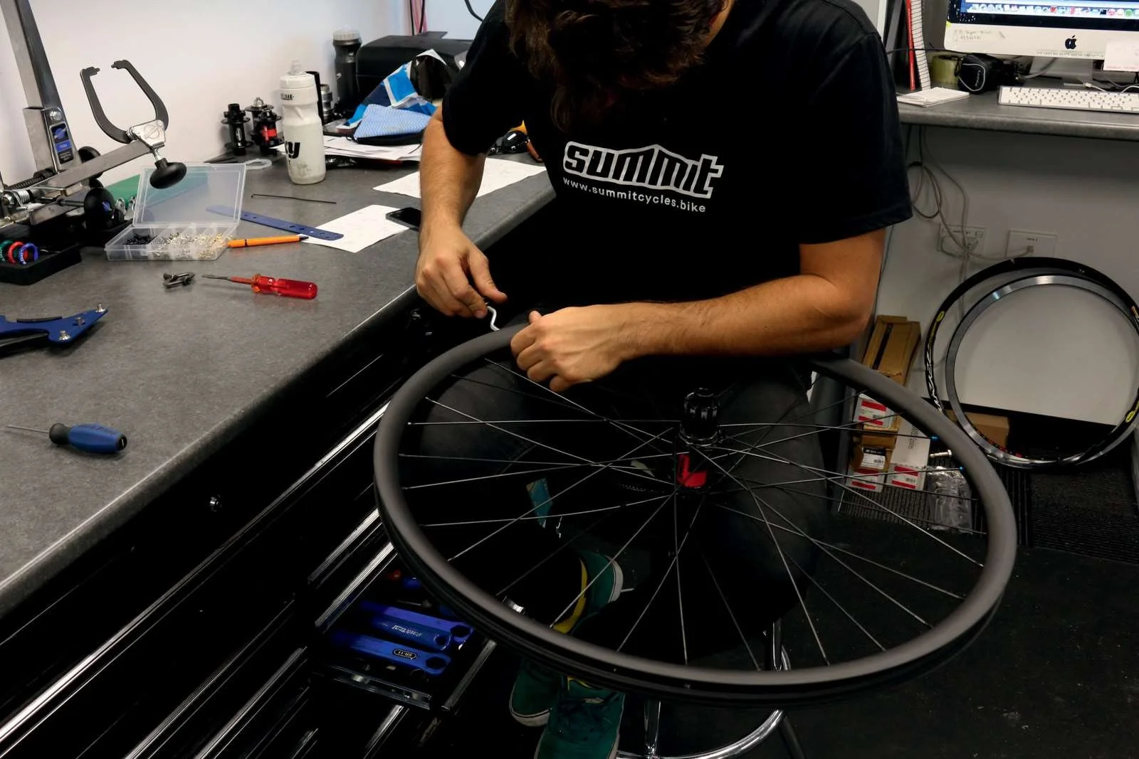 A person working on a bicycle wheel in a workshop, surrounded by tools and equipment.