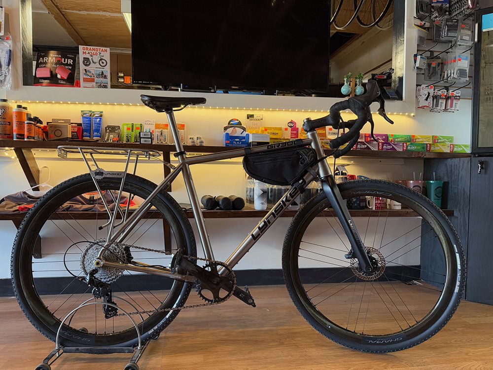 A silver Banshee mountain bike with front suspension, disc brakes, and a black saddle, displayed indoors on a bike stand.