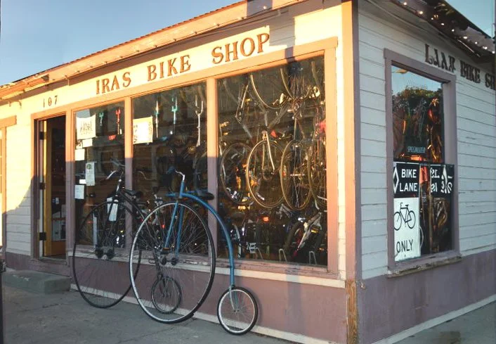 Front of a bicycle shop named Iras Bike Shop with bicycles displayed inside and outside.