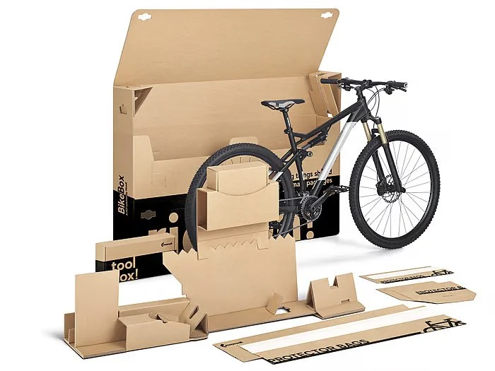 Unassembled black and gray mountain bike with a white suspension fork, surrounded by cardboard packaging and assembly components on a white background.