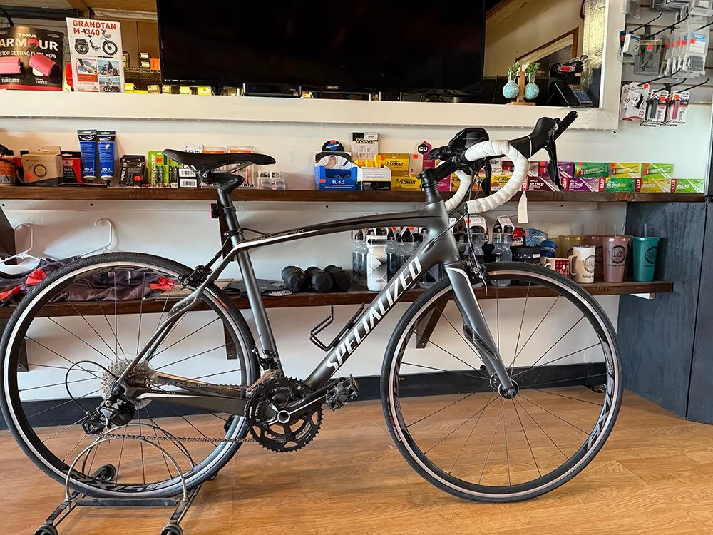 A black Specialized road bicycle with white handlebar tape displayed indoors against a background of various bike accessories and supplies on shelves.
