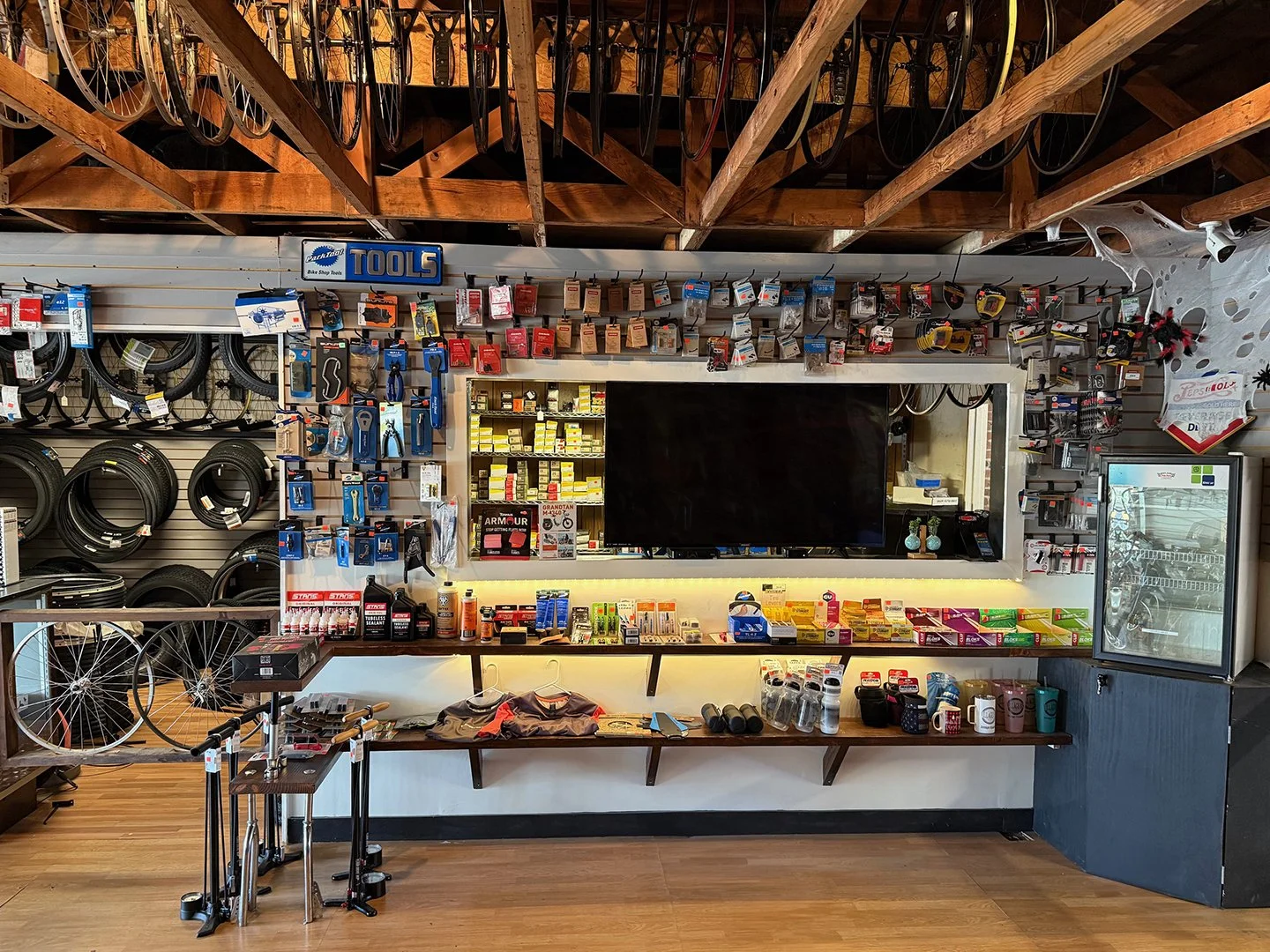 Bike shop with tires, tools, and cycling accessories on display, a large black TV in the center, shelves with various small items, and a refrigerator on the right.