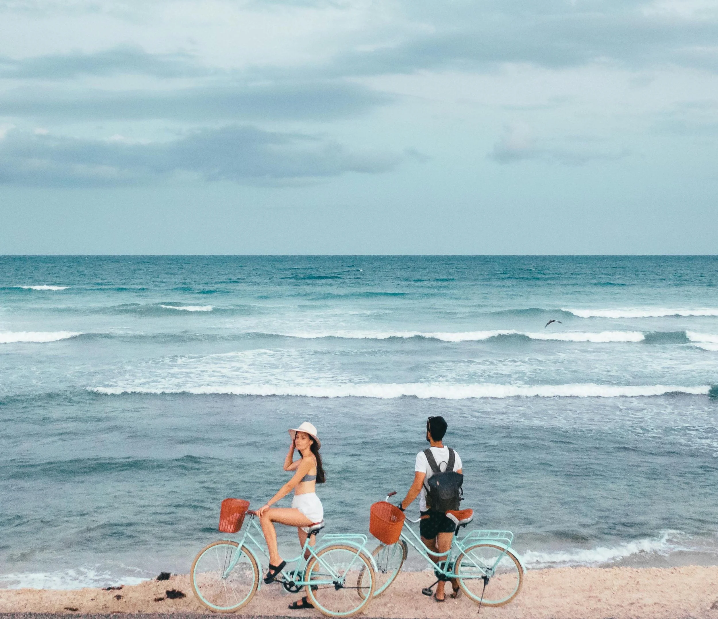 A woman and a man are sitting on bicycles on the beach near the ocean, with waves and cloudy sky in the background.
