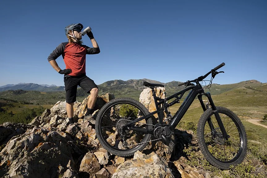 A person wearing a helmet and sunglasses, standing on a rocky hilltop, drinking from a water bottle, with a mountain bike resting on the rocks nearby, under a clear blue sky.