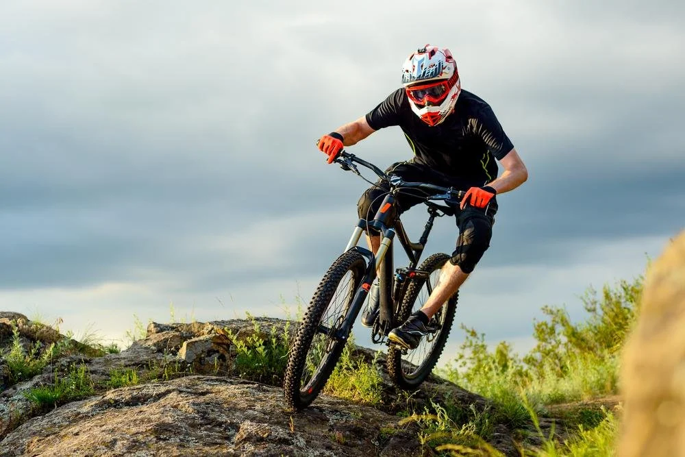 A mountain biker wearing a helmet and gloves riding downhill on a rocky trail with green vegetation and cloudy sky.