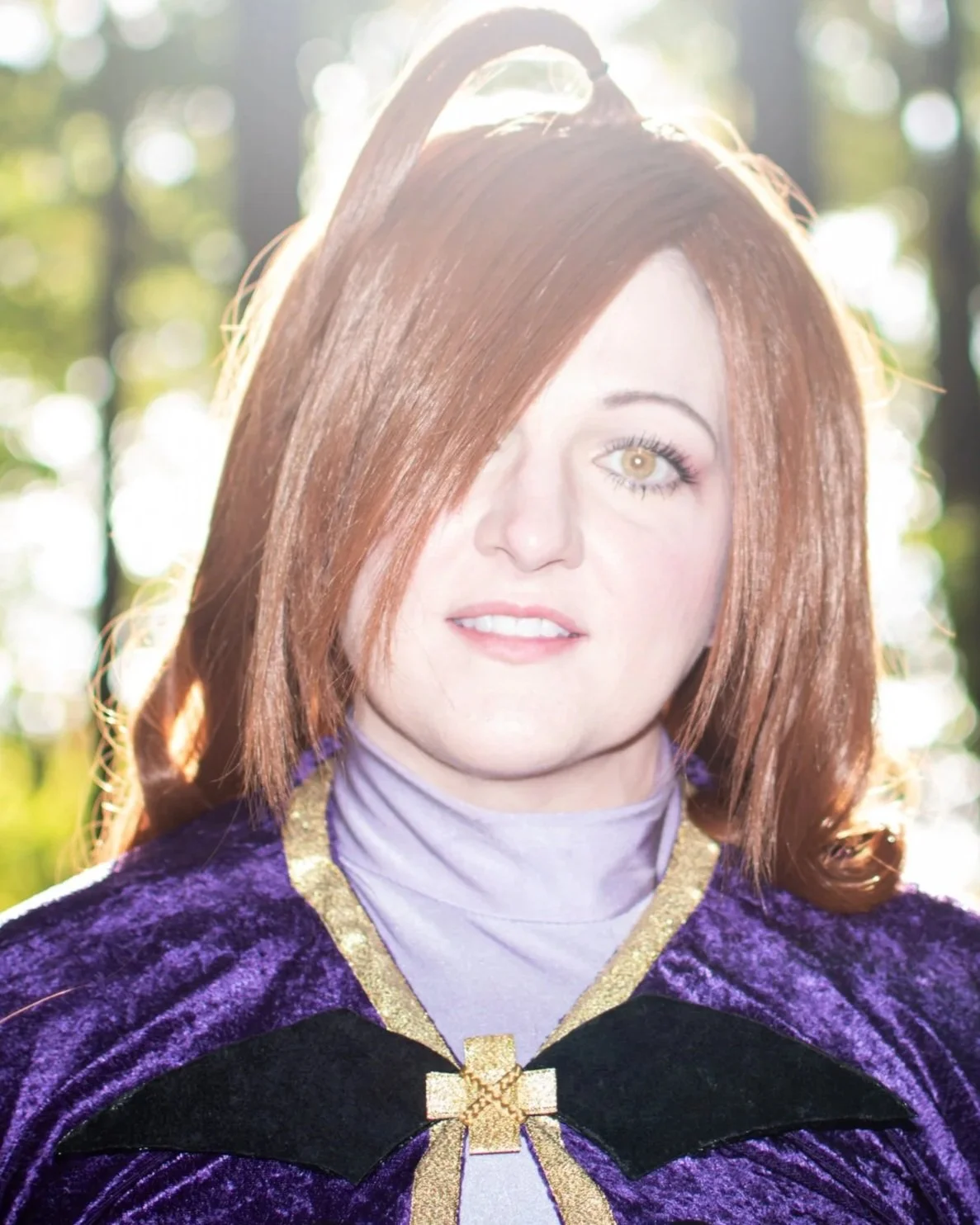 A woman dressed in a costume with a purple velvet cloak, gold trim, and a gold cross pin, outdoors with sunlight filtering through trees in the background.