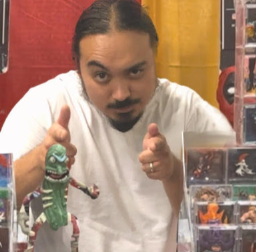 Man with slicked-back hair and mustache pointing at camera, standing behind toy display with Monster High and other dolls.